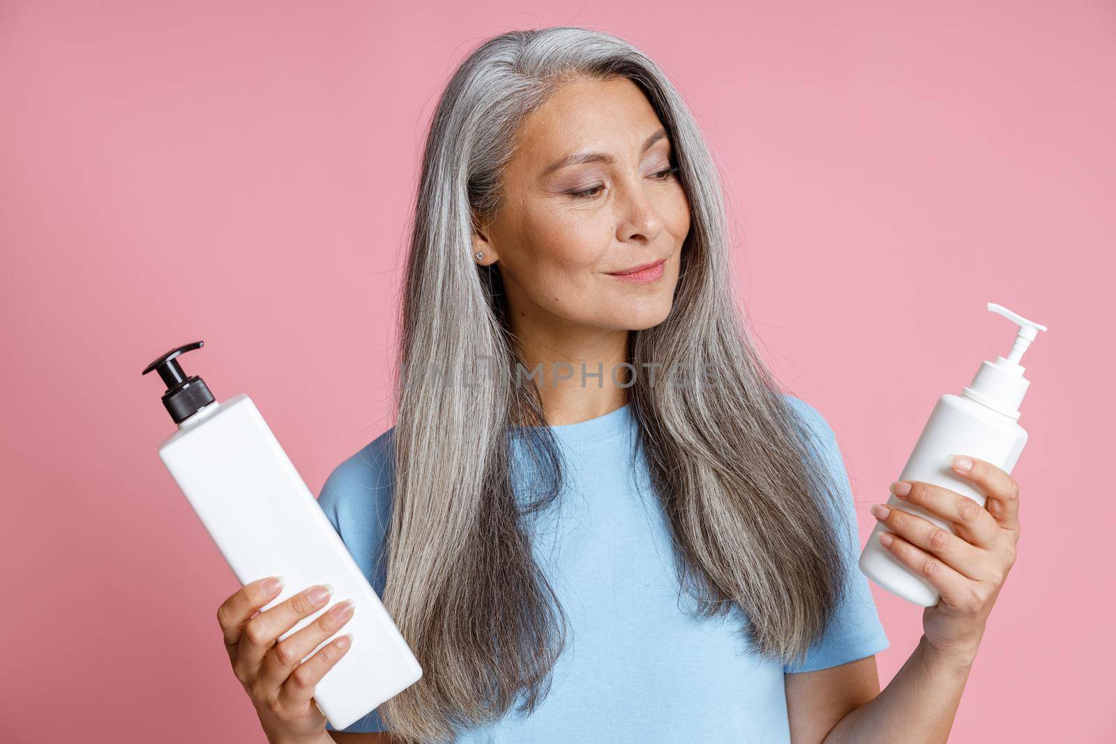 Positive Asian woman in blue t-shirt chooses best hair care product on pink background in studio. Mature beauty lifestyle