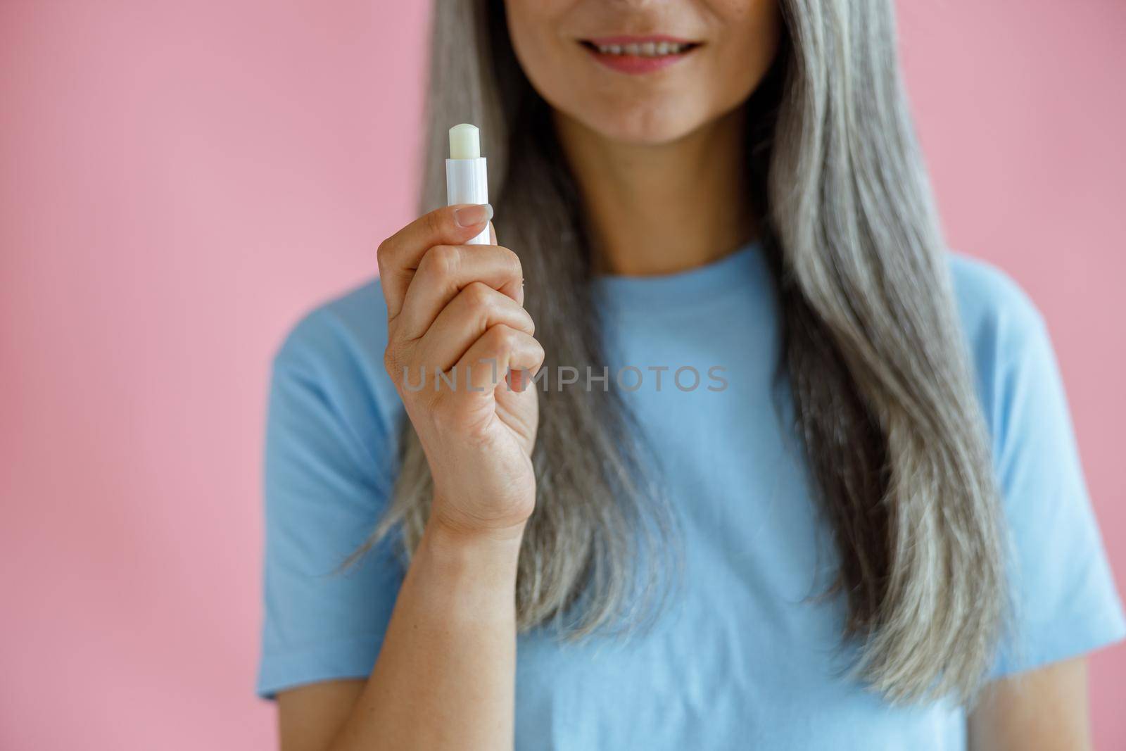 Mature grey haired woman on pink background closeup, focus on hand with lip balm by Yaroslav_astakhov