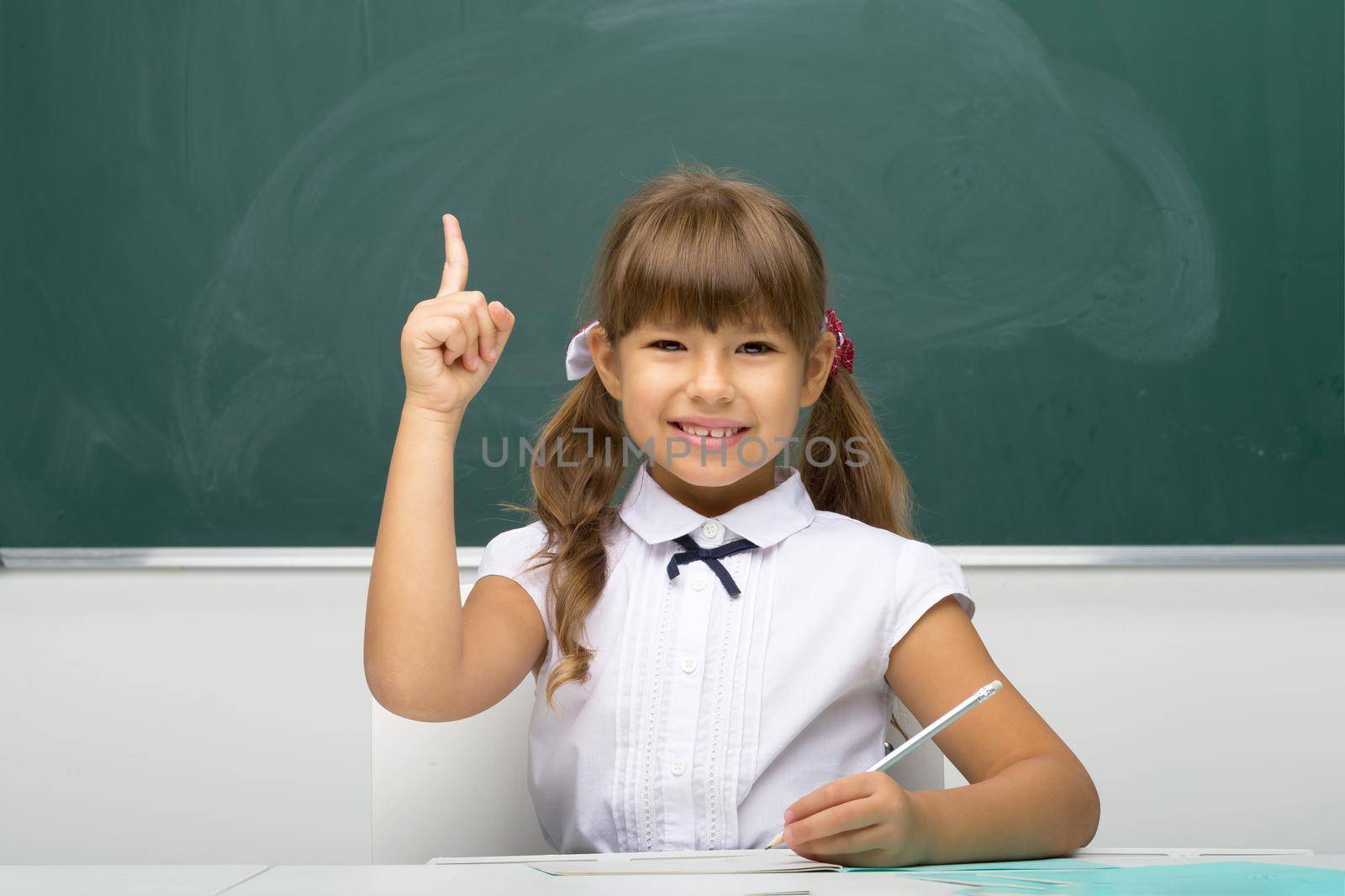 Cheerful schoolgirl pointing up her finger. Pretty girl student in white blouse sitting at desk on background of blackboard in classroom. School and education concept