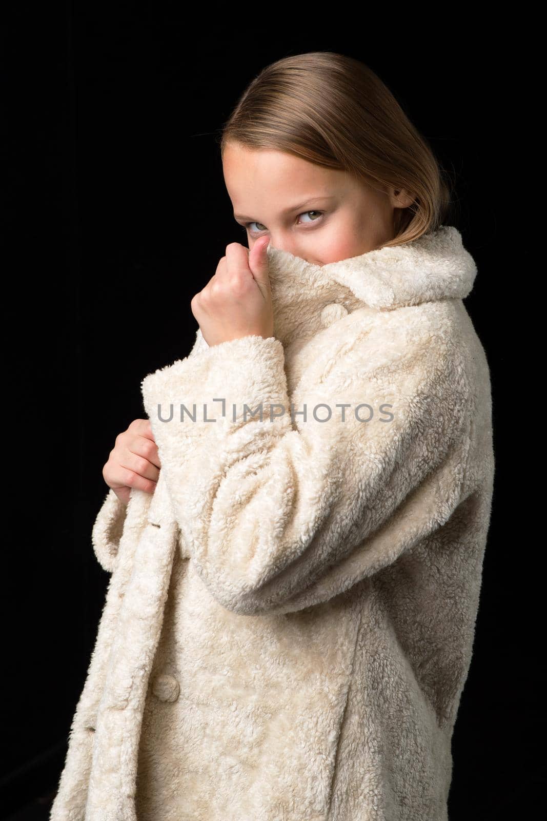 Stylish preteen girl in faux fur coat. Pretty fashionable blonde girl dressed winter coat posing against black background. Close up portrait of fashionable child dressed fashion outwear
