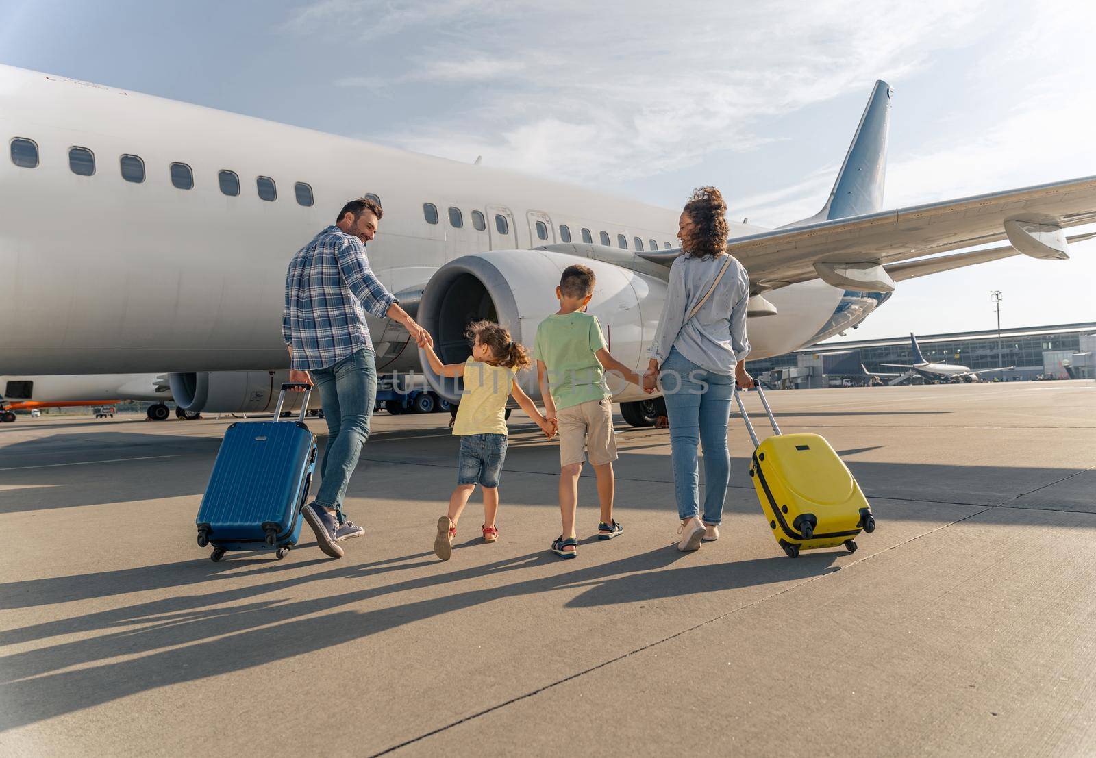 Smiling parents and kids walking with travel bags with a big plane in the background. Trip concept