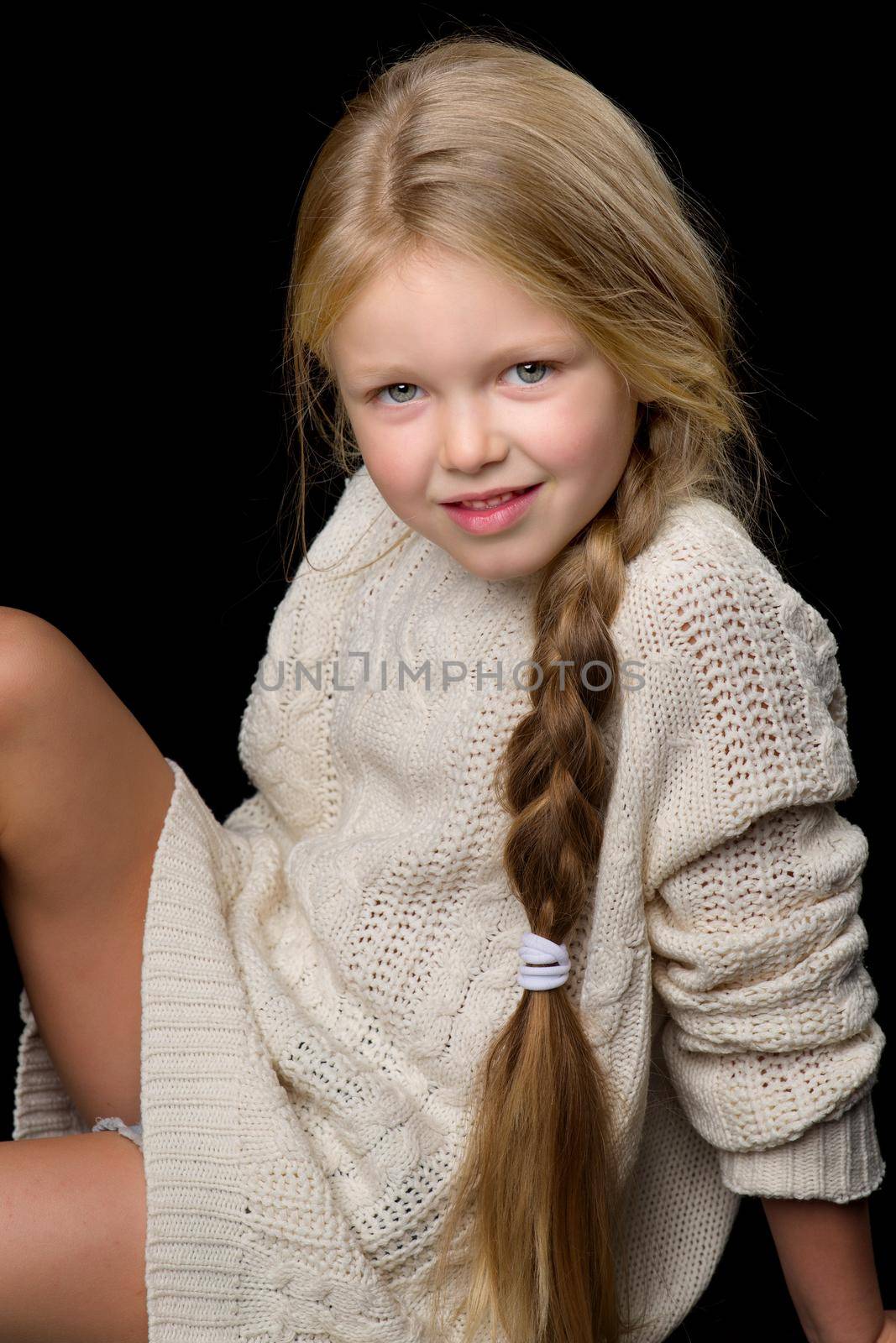 Close up portrait of beautiful girl with braid. Smiling cute little girl wearing warm knitted jumper sitting on floor against black background in studio