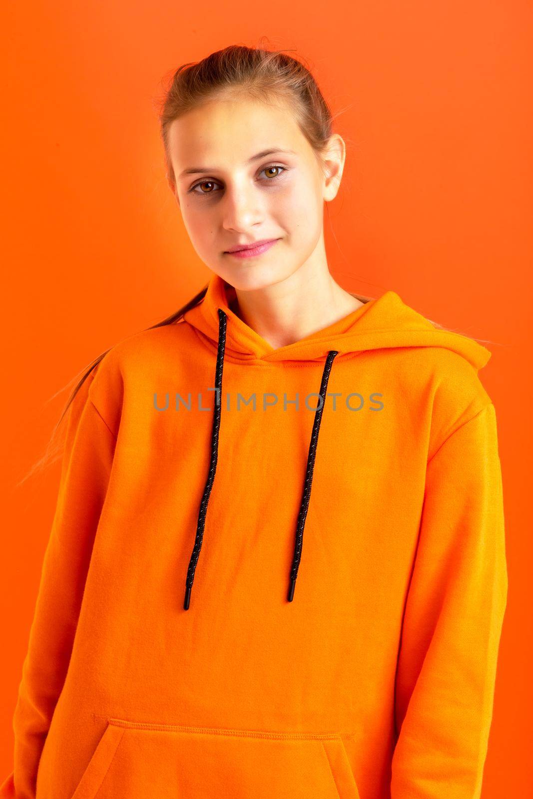 Stylish teenage girl in orange hoodie. Cheerful teenager wearing oversized sweatshirt standing against orange background. Beautiful girl staring happily at camera