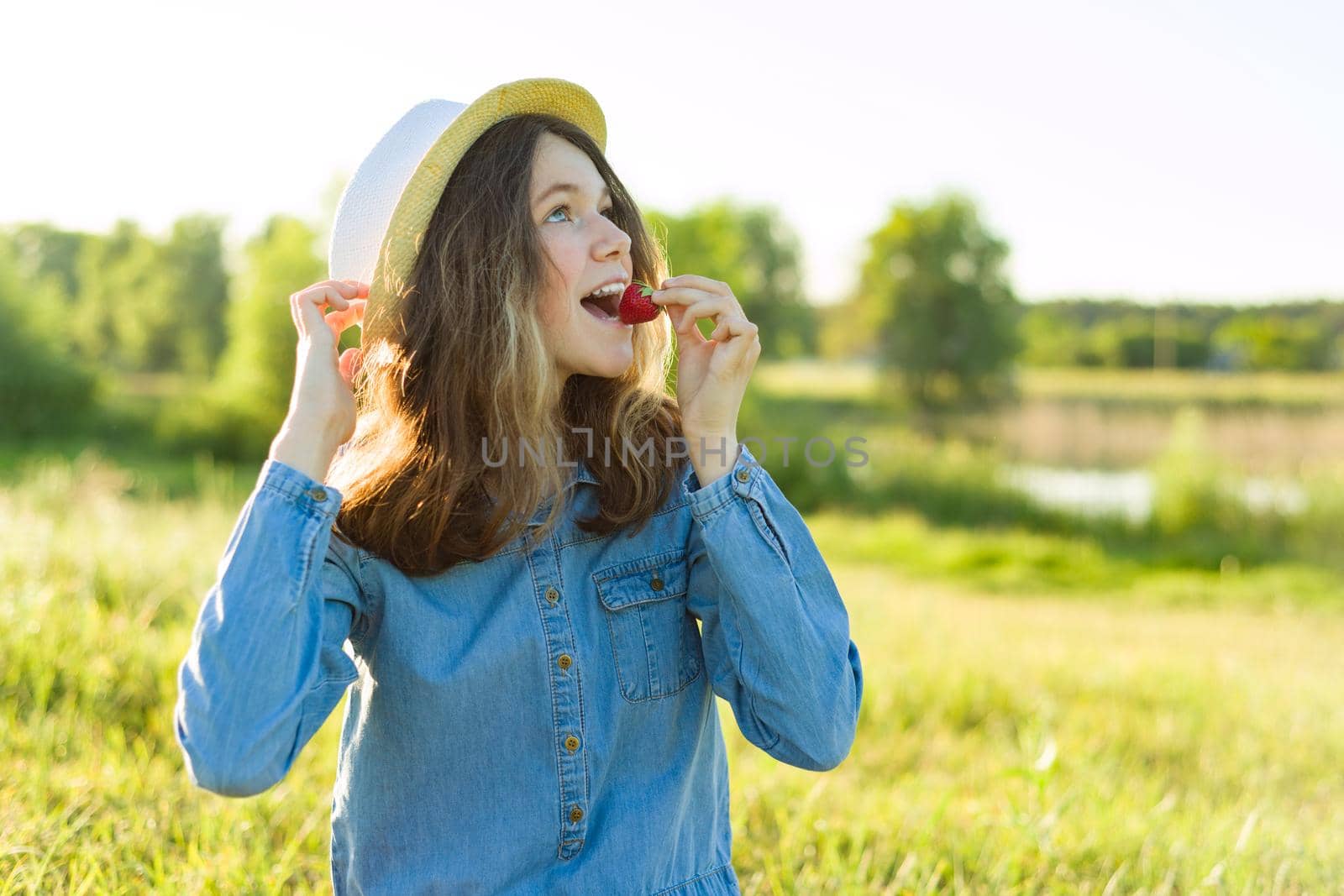 Attractive teen girl eating strawberry. Nature background, rural landscape, green meadow, country style.