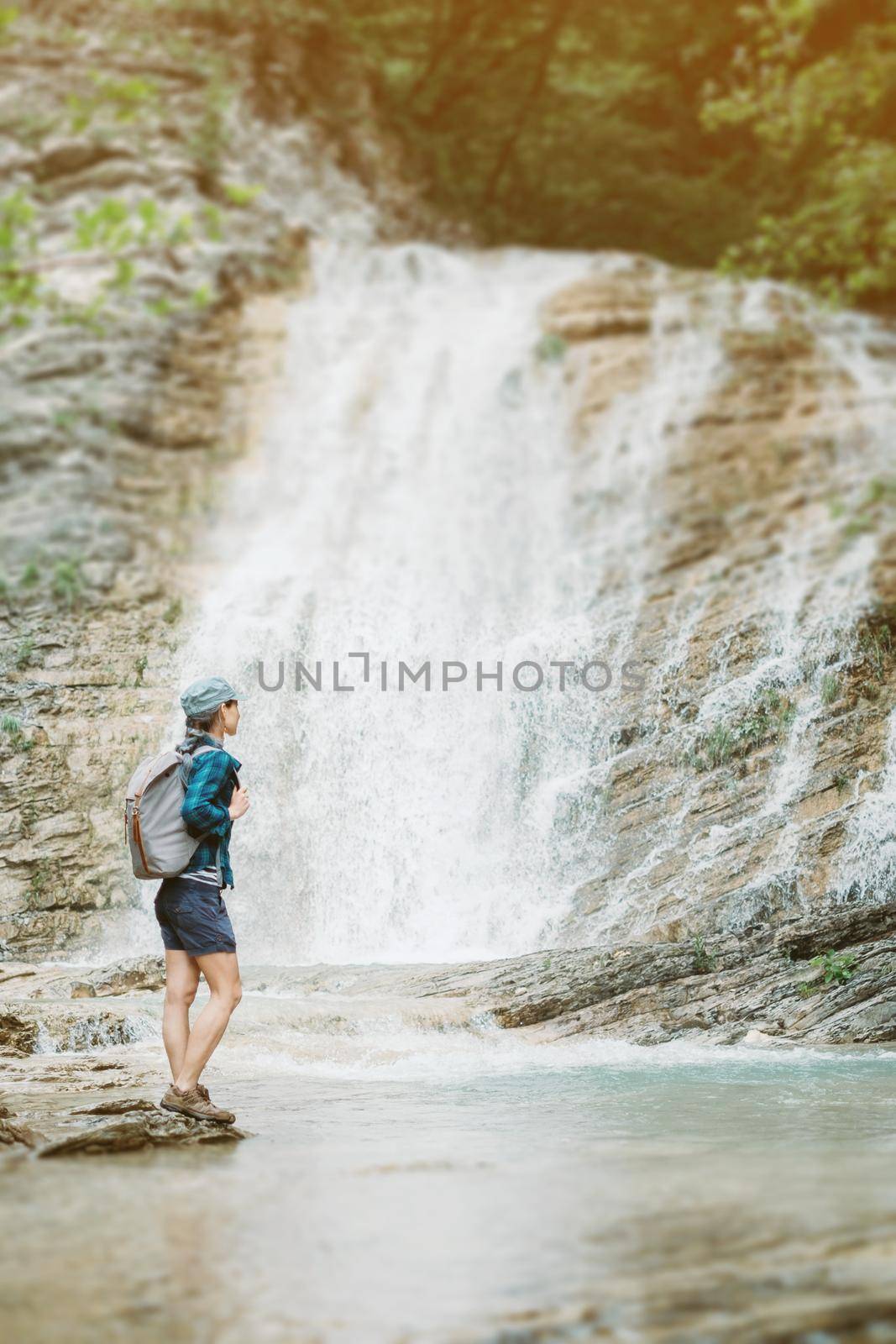 Traveler young woman with backpack standing on bank of river and looking at waterfall in summer on sunny day.