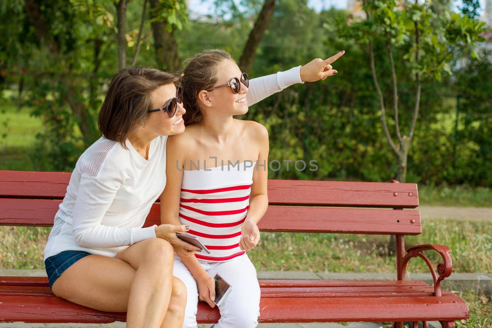 Communication between parent and child. Mom and daughter teenager talking and laughing while sitting on the bench in the park, Mother points with her finger to the side