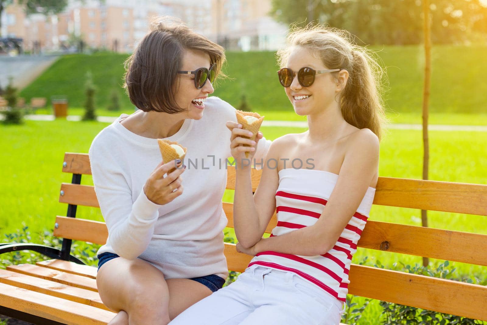 Holiday with the family. Happy young mother and cute daughter teenager in city park eating ice cream, talking and laughing.