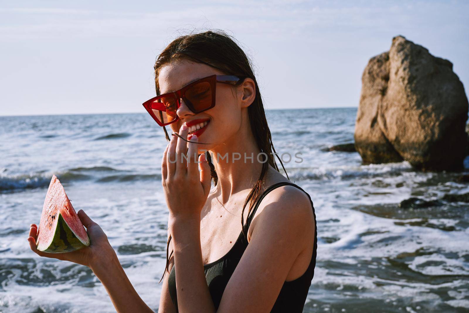 cheerful woman in black swimsuit eating watermelon on the beach by Vichizh