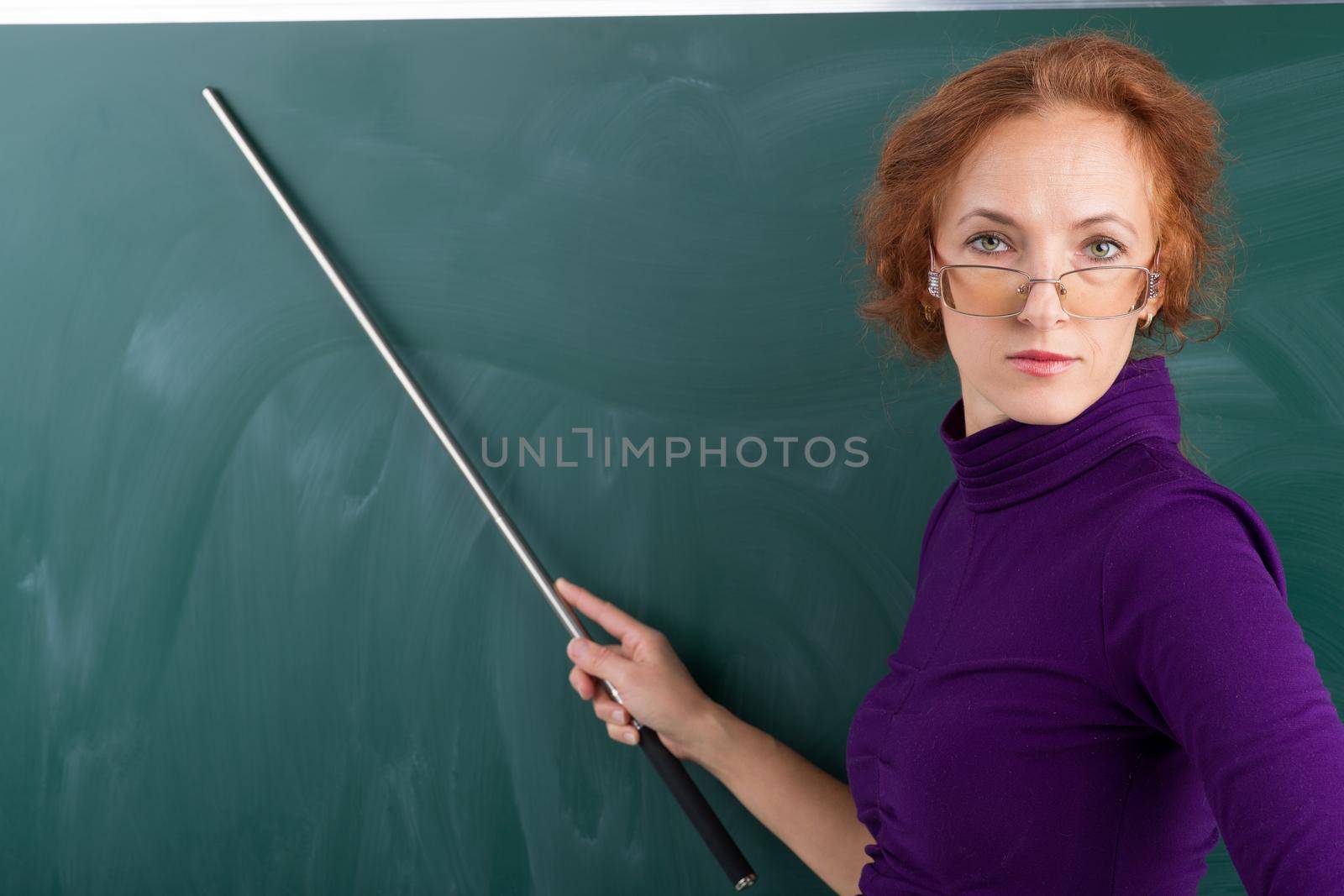 Teacher standing in front of blackboard. Young woman wearing glasses and casual dress standing with wooden pointer and looking down. Teacher posing at green chalkboard and pointing at something