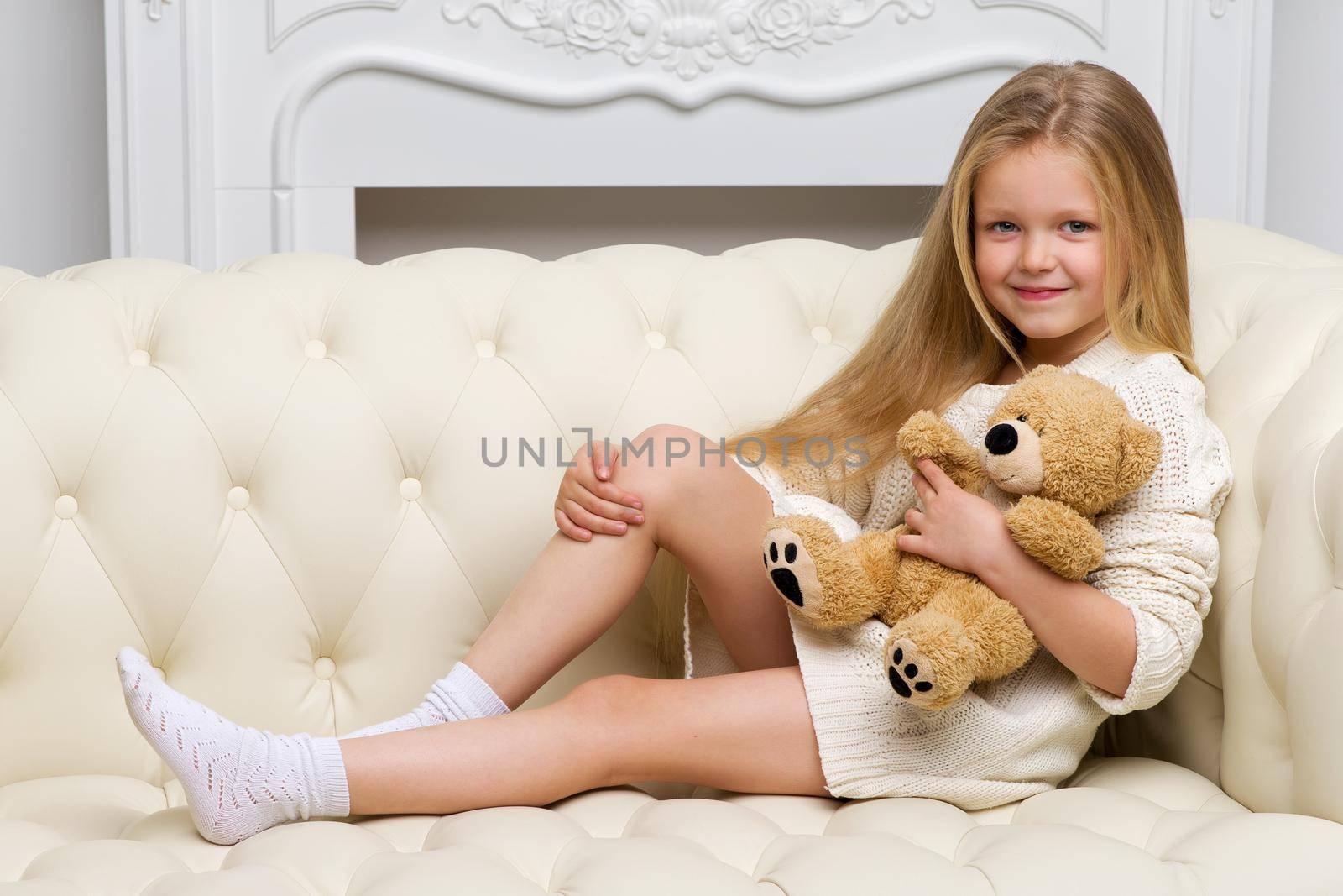 Lovely blonde girl sitting on comfortable sofa hugging teddy bear. Portrait of cute long haired girl wearing warm knitted dress. Adorable happy child resting on couch with favorite stuffed toy