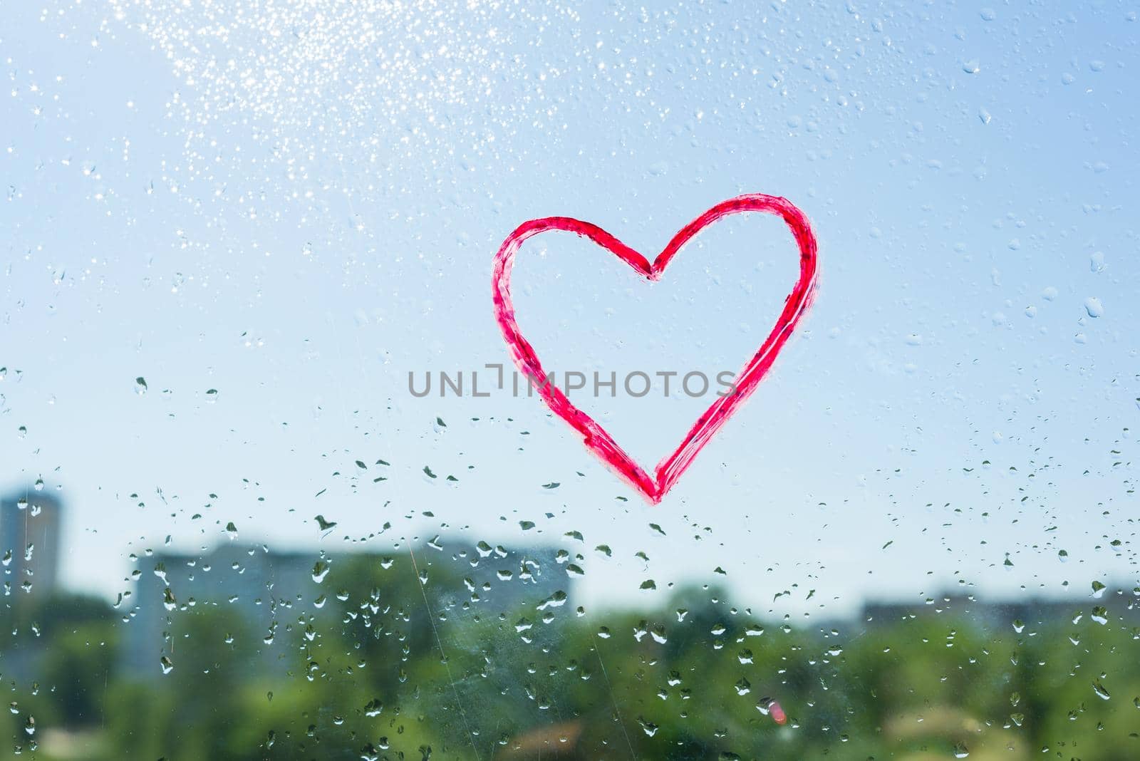 Red heart painted with lipstick on the window with water drops. Background blue sunny sky, drops shine in the sun. by VH-studio