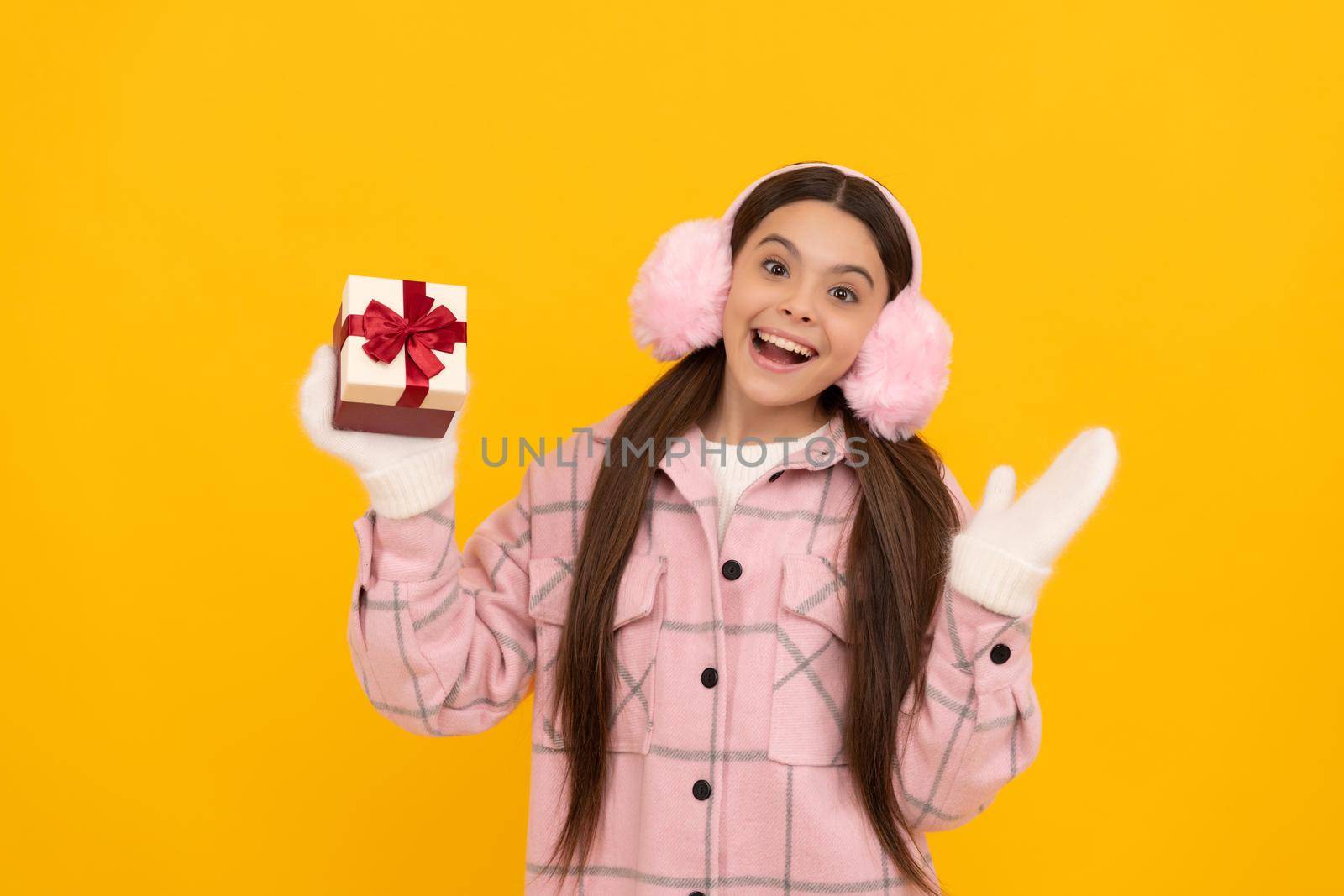 teen girl in mittens hold present on yellow background. xmas holiday gift. child wearing warm clothes. boxing day. happy new year. merry christmas. happy amazed kid in winter earmuffs with box.