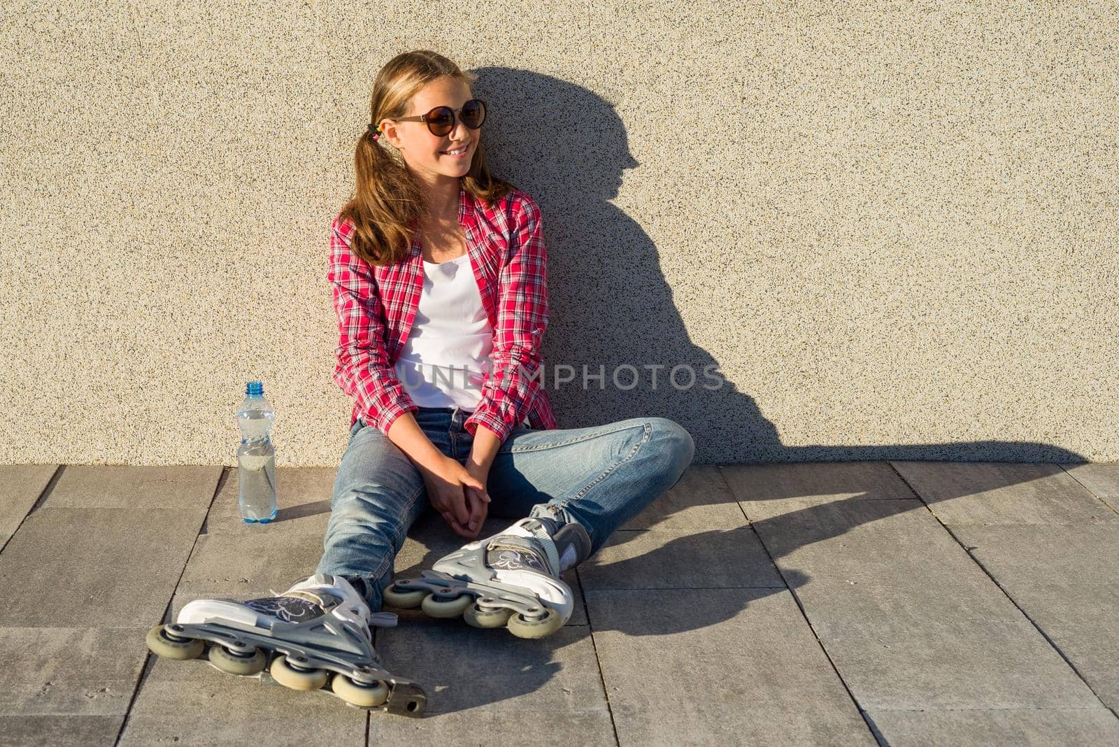 Young smiling cool girl shod in rollerblades, sits on the sidewalk and holding a water bottle. by VH-studio