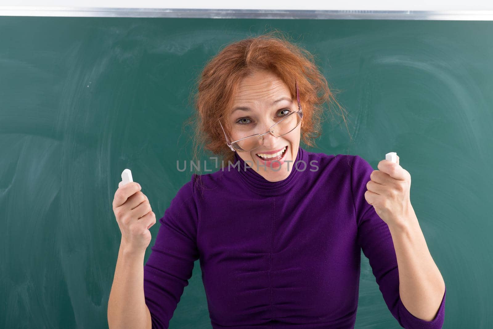 Teacher holding pieces of chalks in her hands. Excited young woman wearing glasses and casual dress standing in front of blackboard. Cheerful teacher posing at green chalkboard