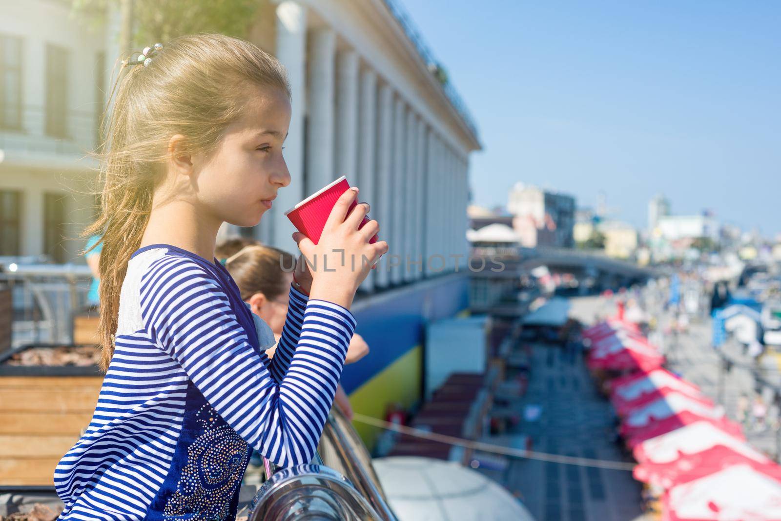 Portrait of a cool girl 10 years old, in profile, drinks from a glass and looks afar on the river. City Embankment, European city