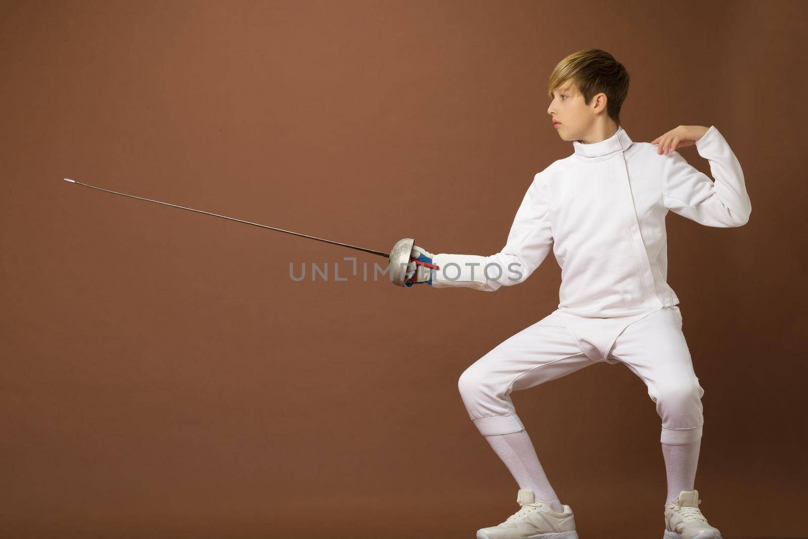 Boy fencer standing in attacking pose. Portrait of teenage boy wearing white fencing suit posing with sabre against brown background. Teenager taking part at foil fencing tournament