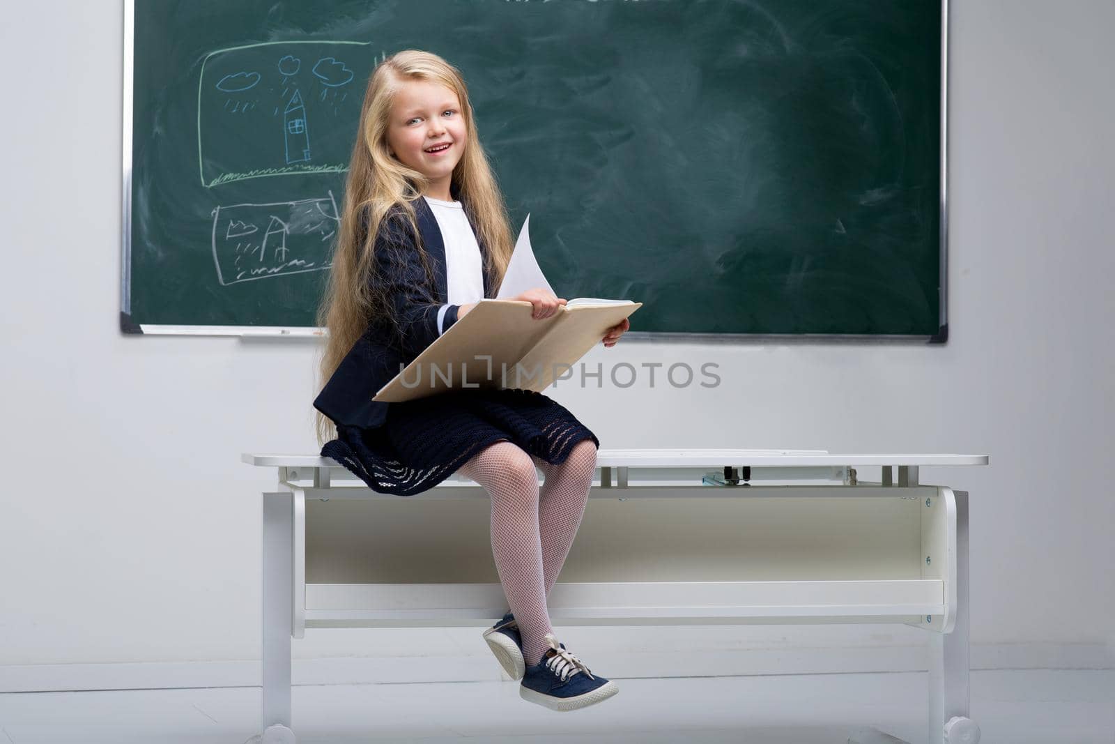 Beautiful little schoolgirl sitting on desk. Blonde elementary school student posing on desk with book on background of blackboard in classroom. Back to school, education concept