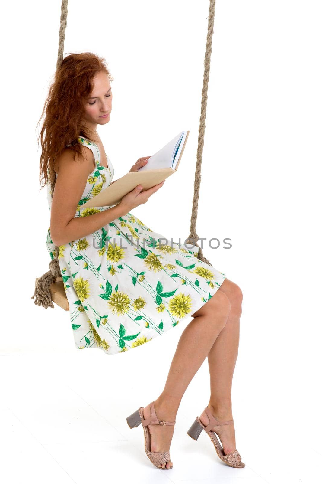 Happy young woman reading book on swing. Smiling woman wearing summer dress sitting on rope swing against white background. Portrait of beautiful girl posing in studio