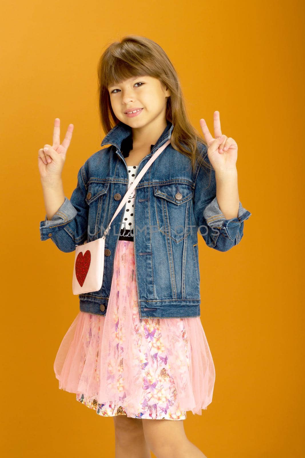 Beautiful girl showing victory signs. Cheerful emotional preteen girl wearing denim jacket and fluffy skirt showing peace gesture raised two hands against ocher background