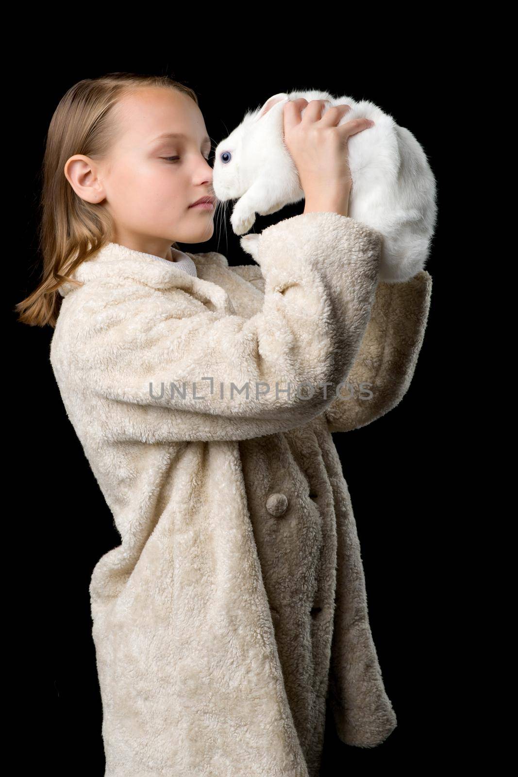 Pretty stylish girl with white rabbit. Side shot of beautiful girl in beige fur coat looking at cute bunny pet. Portrait of preteen child posing in studio against black background