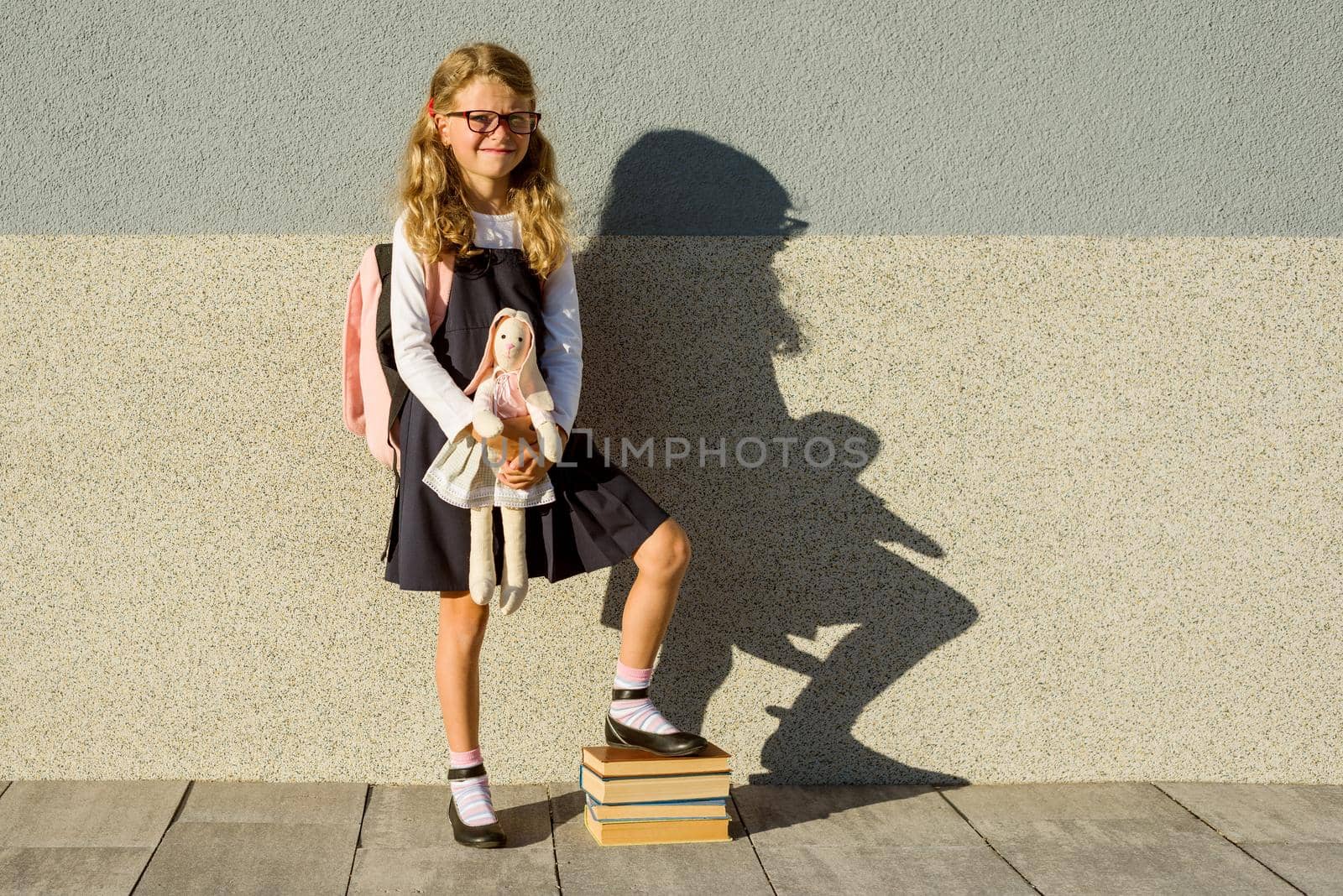 A cute schoolgirl of primary school is smiling with a toy in her hand. Girl with a backpack near the building outdoors. Leaving childhood and Back to school!