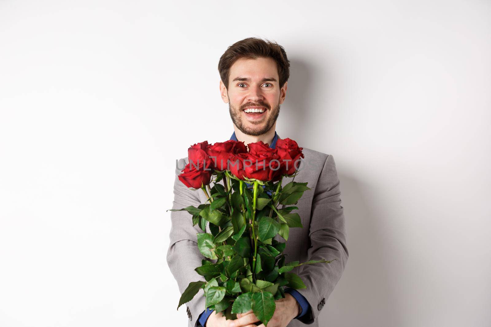 Handsome man in love wishing happy valentines day, giving bouquet of flowers on romantic date, smiling at camera, wearing suit over white background.