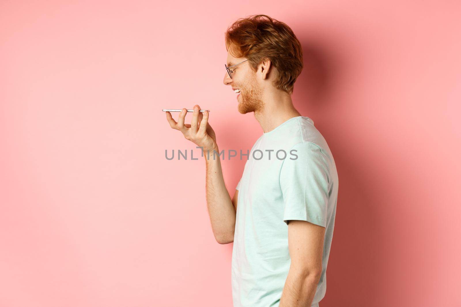 Profile portrait young man with red hair, smiling pleased while record voice message on smartphone, talking to virtual assistant, standing over pink background.
