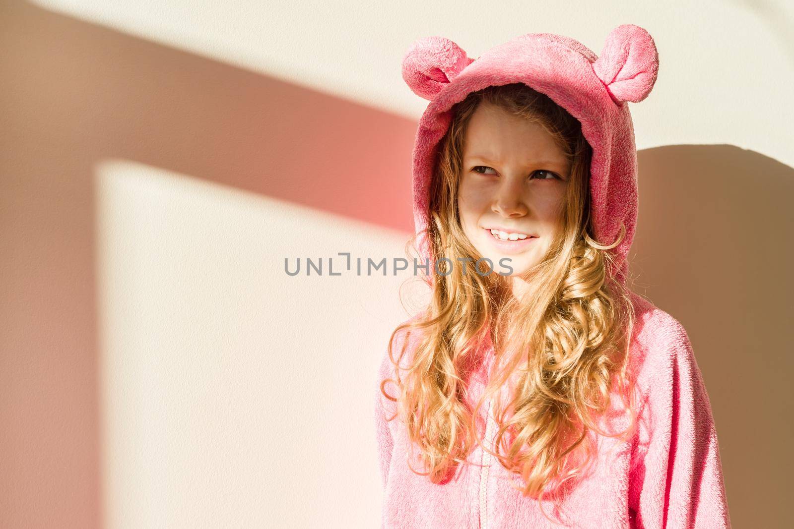 Portrait of a girl in soft warm pink pajamas. Girl 7 years old, blonde with long curly hair, in a hood, looking away. Background light house wall, solar lighting from the window, copy space