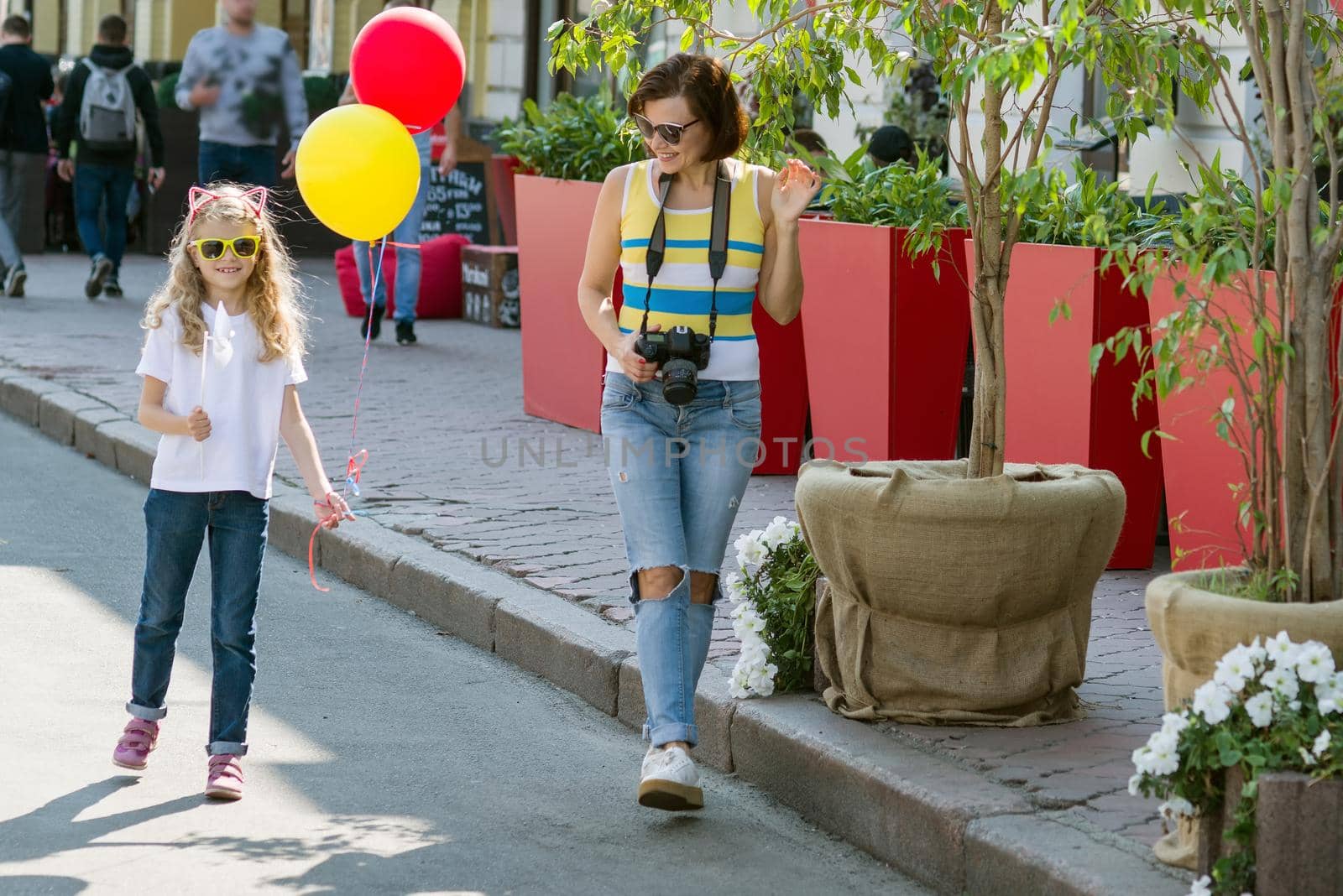 Urban portrait of mother and daughter. Girl with balloons is walking along the city street.