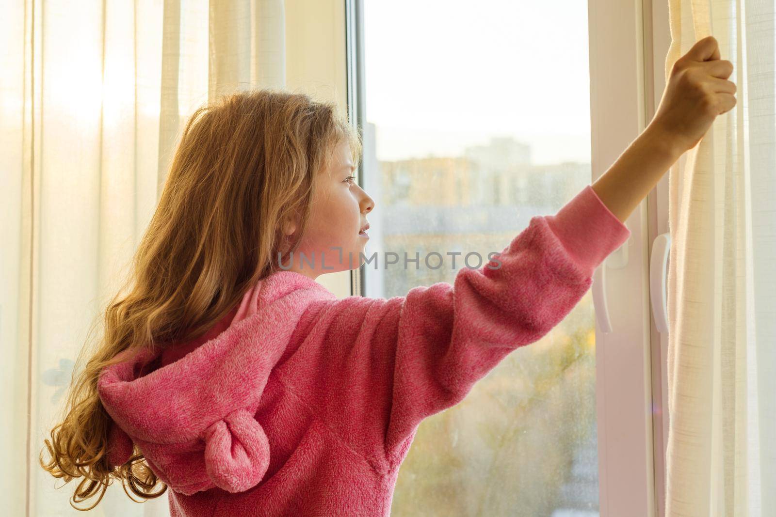 Good morning. Girl child in pajamas opens curtains and looks out the window