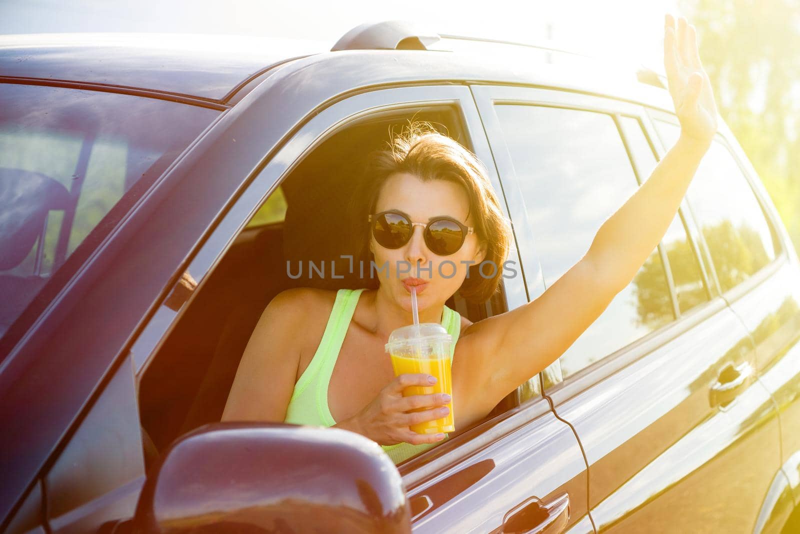 A happy woman driver stops to relax, enjoy the journey, drink orange juice and wave your hand