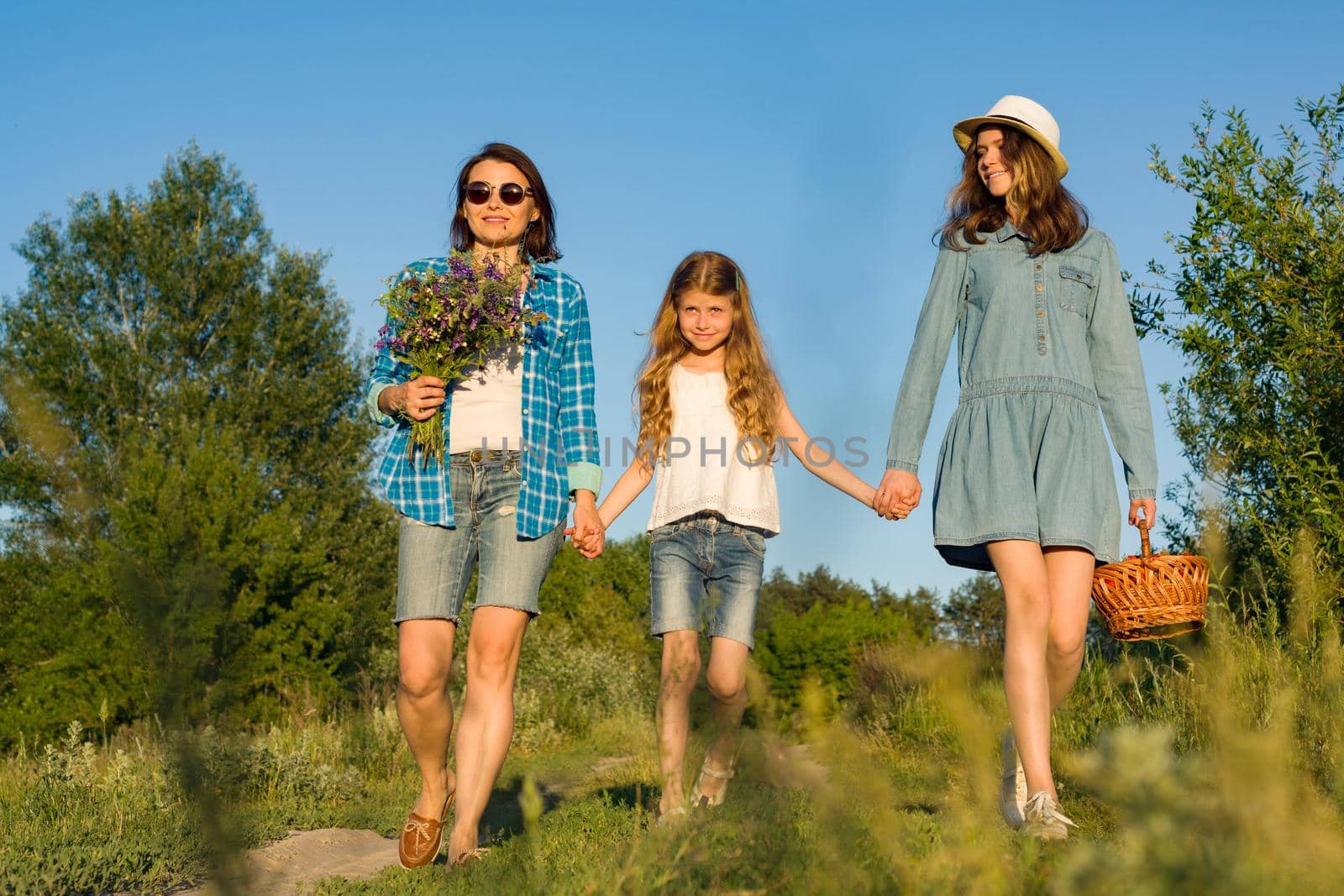 Mother and daughter hold hands, walk along the country road. With wildflowers a basket with berries.