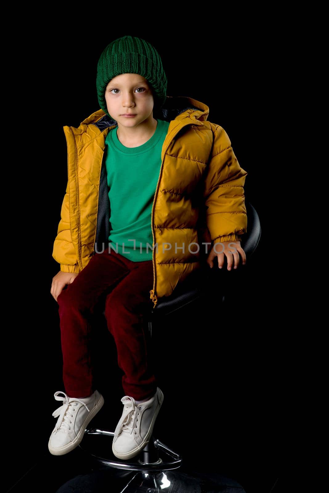 Cute boy in winter jacket and knitted hat. Stylish cheerful boy sitting on chair posing at camera in studio. Child wearing fashionable winter outerwear isolated on black background