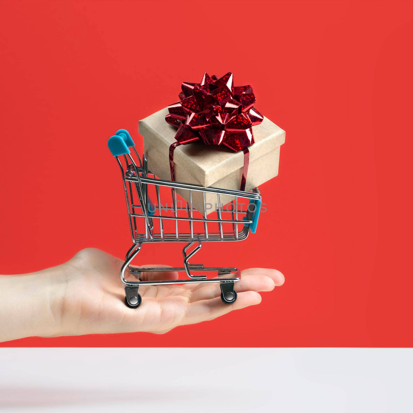 Hand holding Toy shopping cart or trolley with large gift box. A gift for a holiday, anniversary, or birthday. Trolley cart with a gift box Isolated over red background