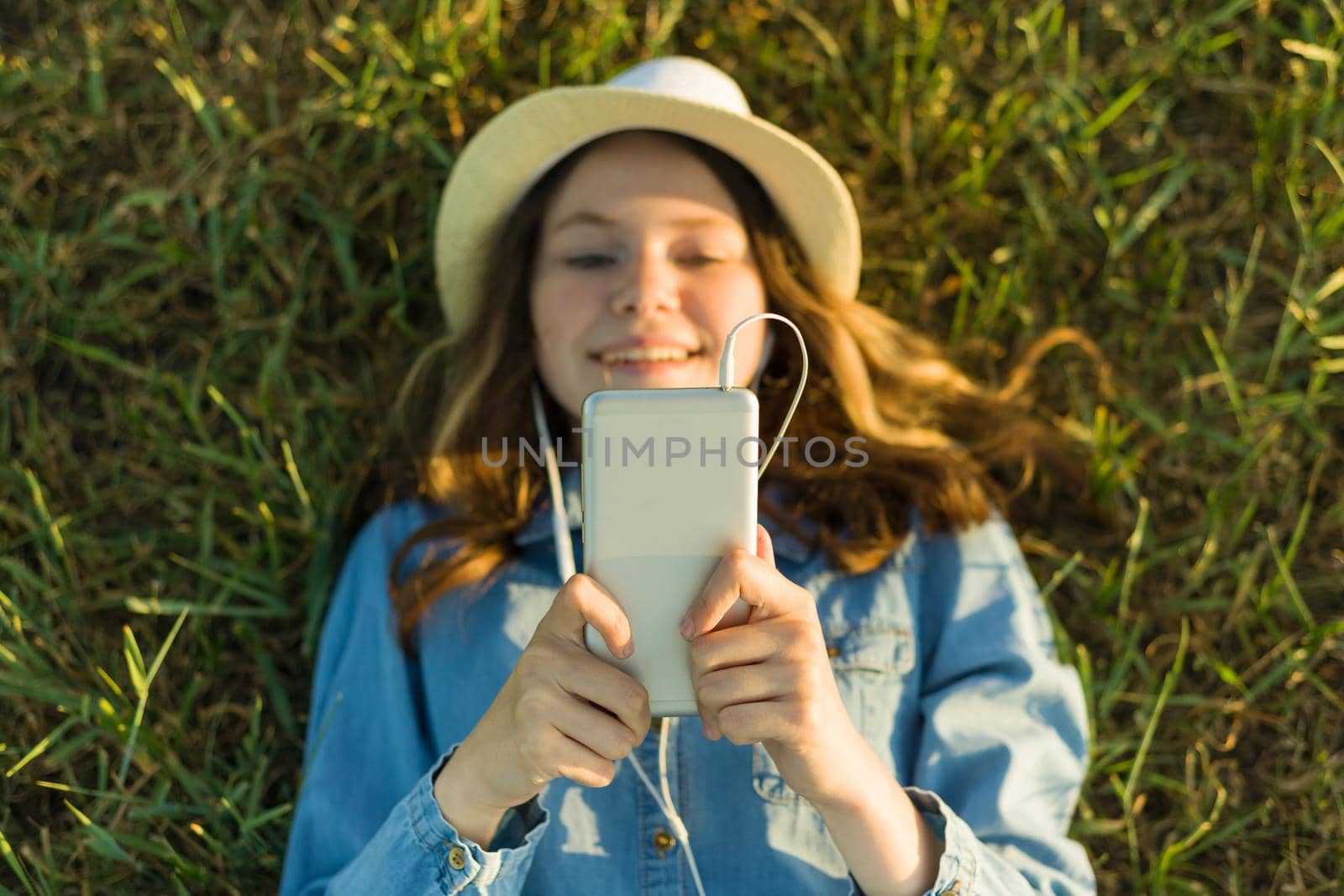 Teenage girl in hat with headphones lies on the green grass and looks into the phone. Focus on smartphone, top view