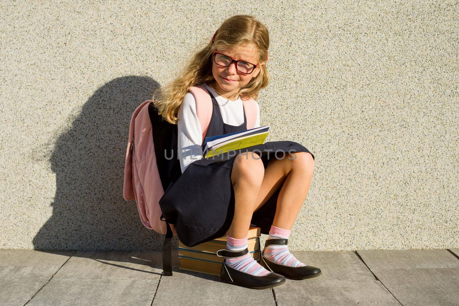 An elementary school student with notebooks in his hand. A little schoolgirl with a backpack near the building in the open air.