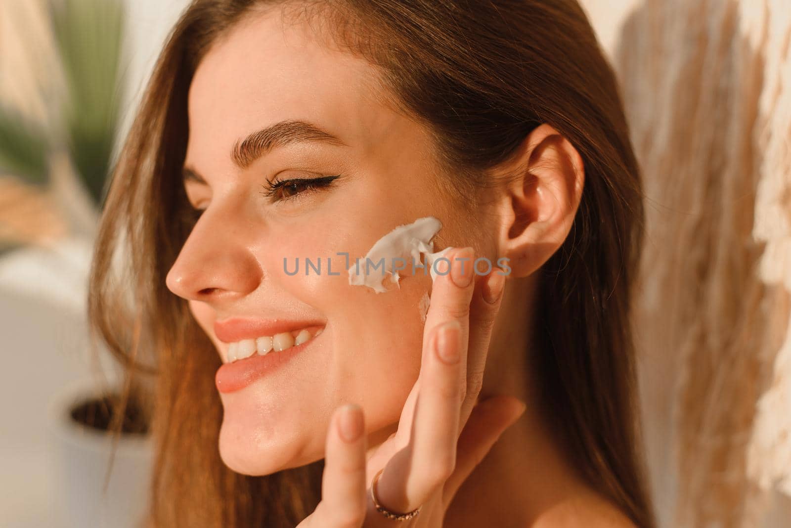 A young woman applies moisturizer to her face in her sun bath. Daily routine, hydration and skin care concept.