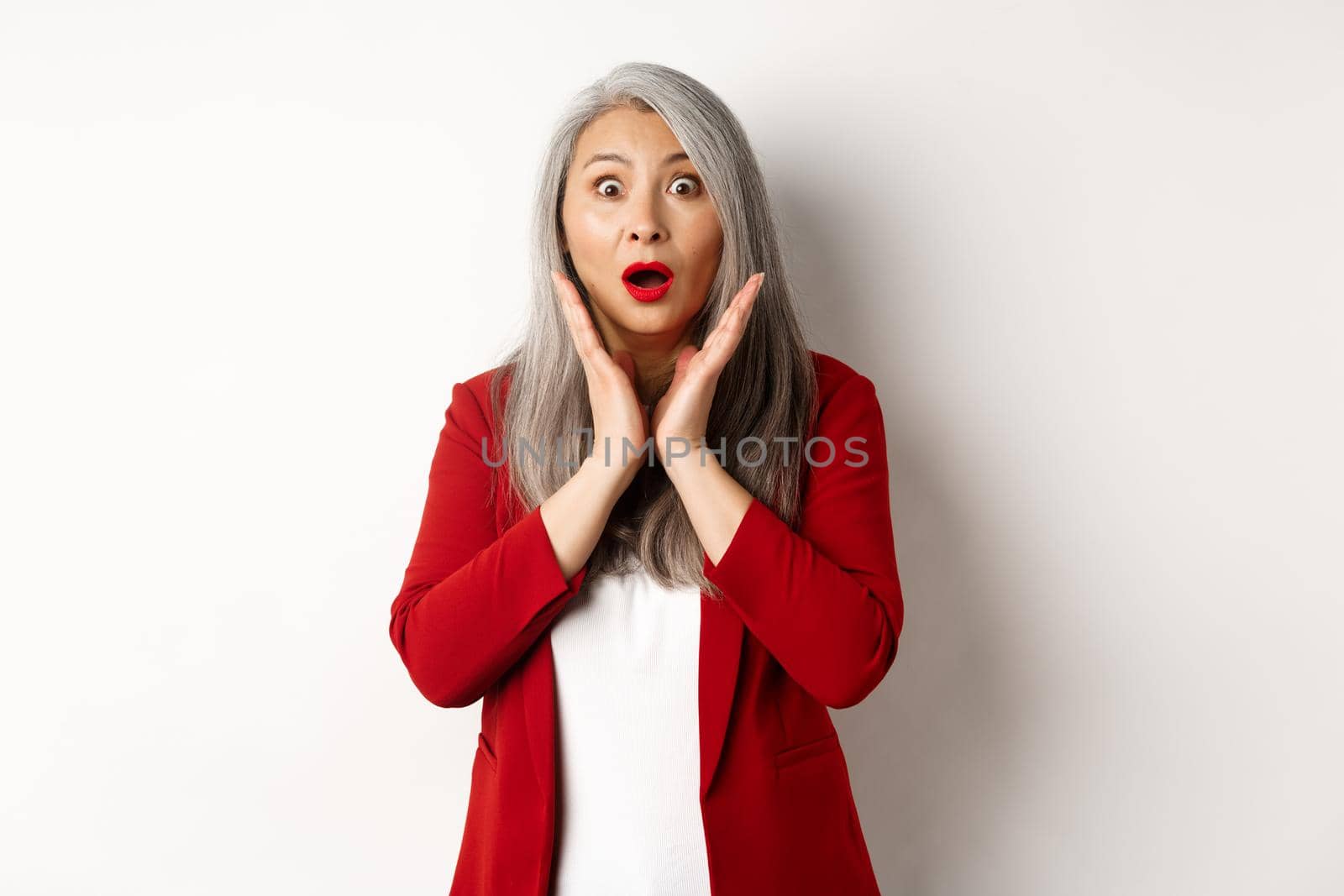 Business people. Asian senior businesswoman in red blazer and makeup gasping, looking surprised at camera, standing over white background.
