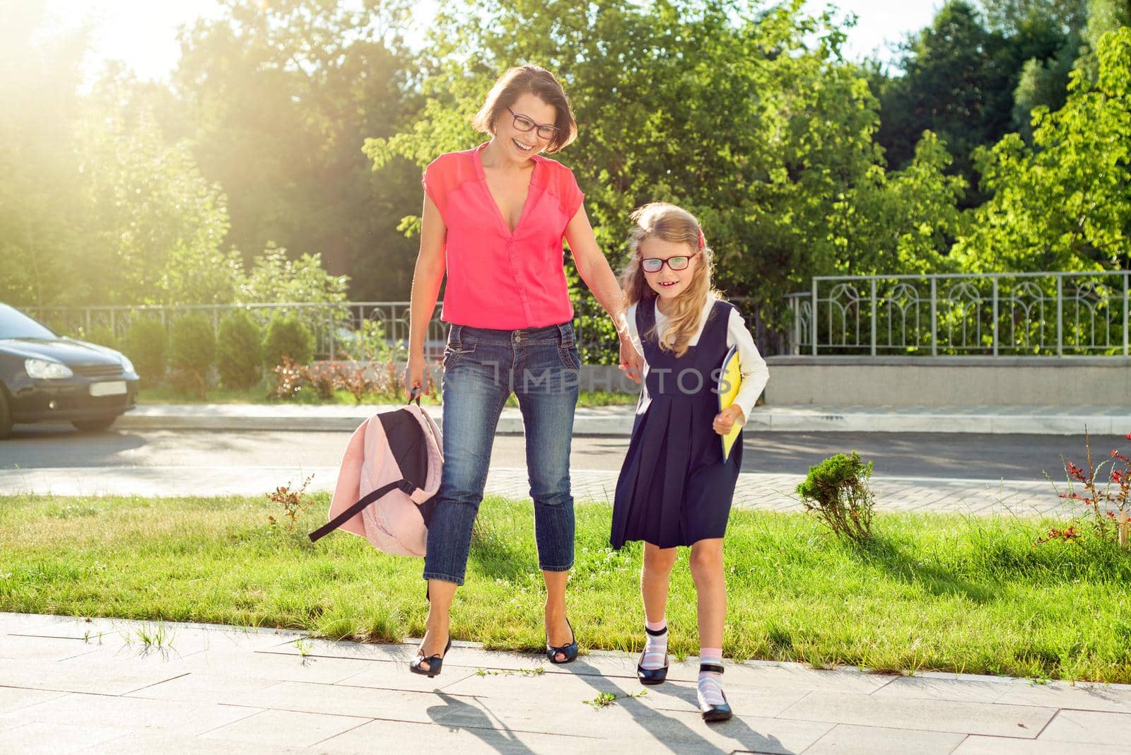 Mom and pupil of elementary school holding hands. The parent takes the child to school. Outdoors, return to the concept of the school.