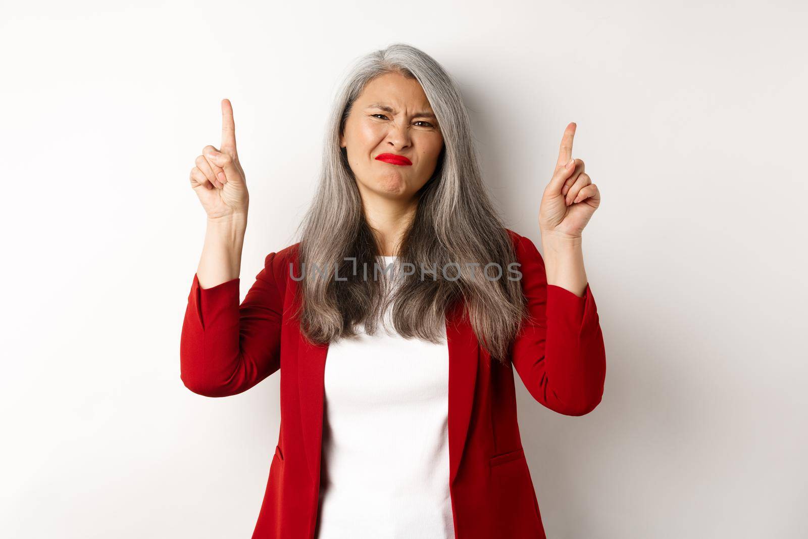 Skeptical and disappointed asian businesswoman pointing fingers up, grimacing and showing bad promo offer, standing over white background.