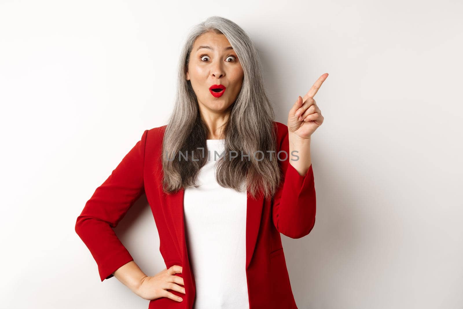 Amazed senior asian lady pointing upper right corner, staring excited at camera, checking out promo offer, standing over white background.