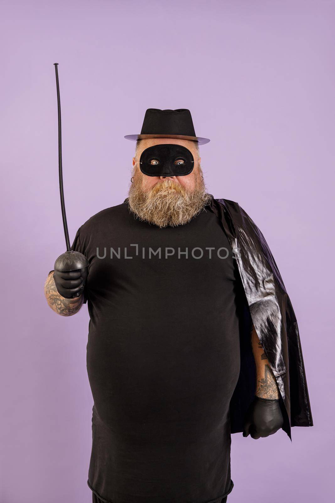 Funny mature bearded male person with overweight wearing Zorro suit with cape, mask and toy epee stands on purple background in studio