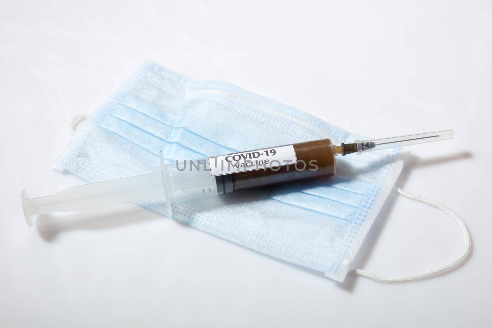 Close up view of needle on a syringe filled with vaccine laying on face mask. on gray background