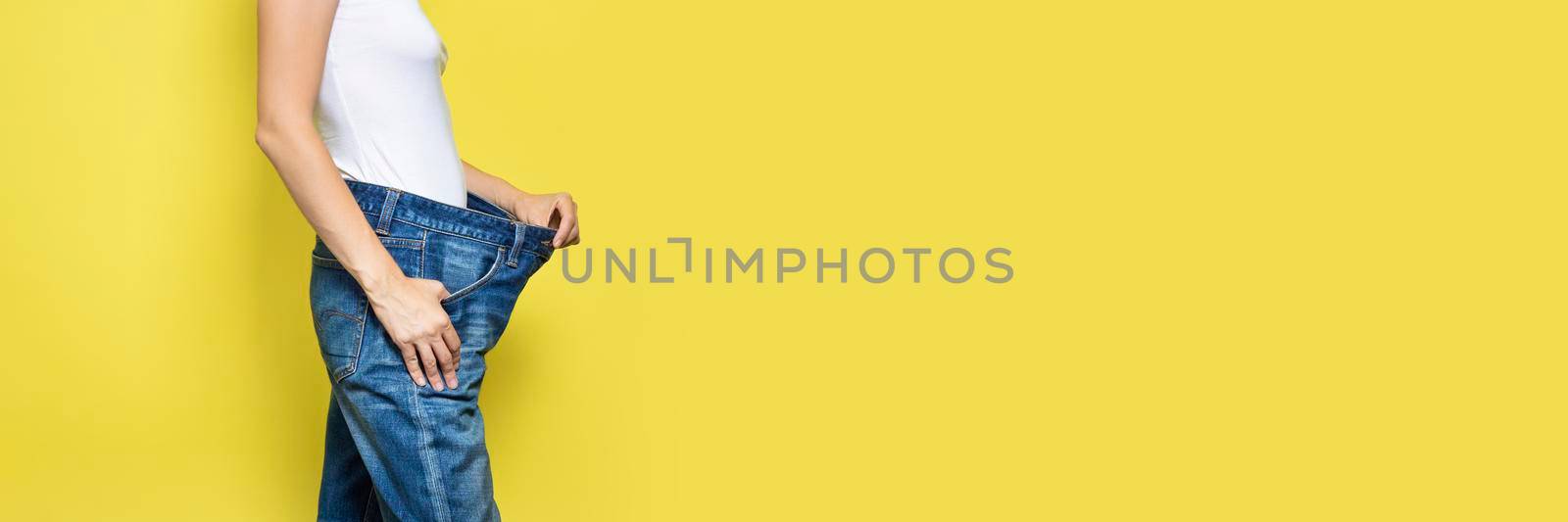 The concept of diet, proper nutrition, weight loss. Slim Woman Showing Loose Jeans and her Loss Weight. Woman white t-shirt and oversize jeans isolated on yellow background. Banner with copy space by esvetleishaya