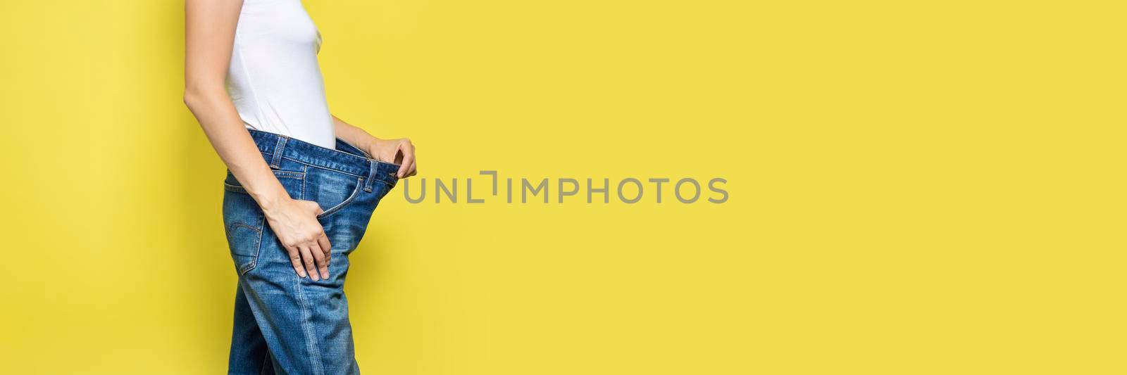 The concept of diet, proper nutrition, weight loss. Slim Woman Showing Loose Jeans and her Loss Weight. Woman white t-shirt and oversize jeans isolated on yellow background. Banner with copy space.