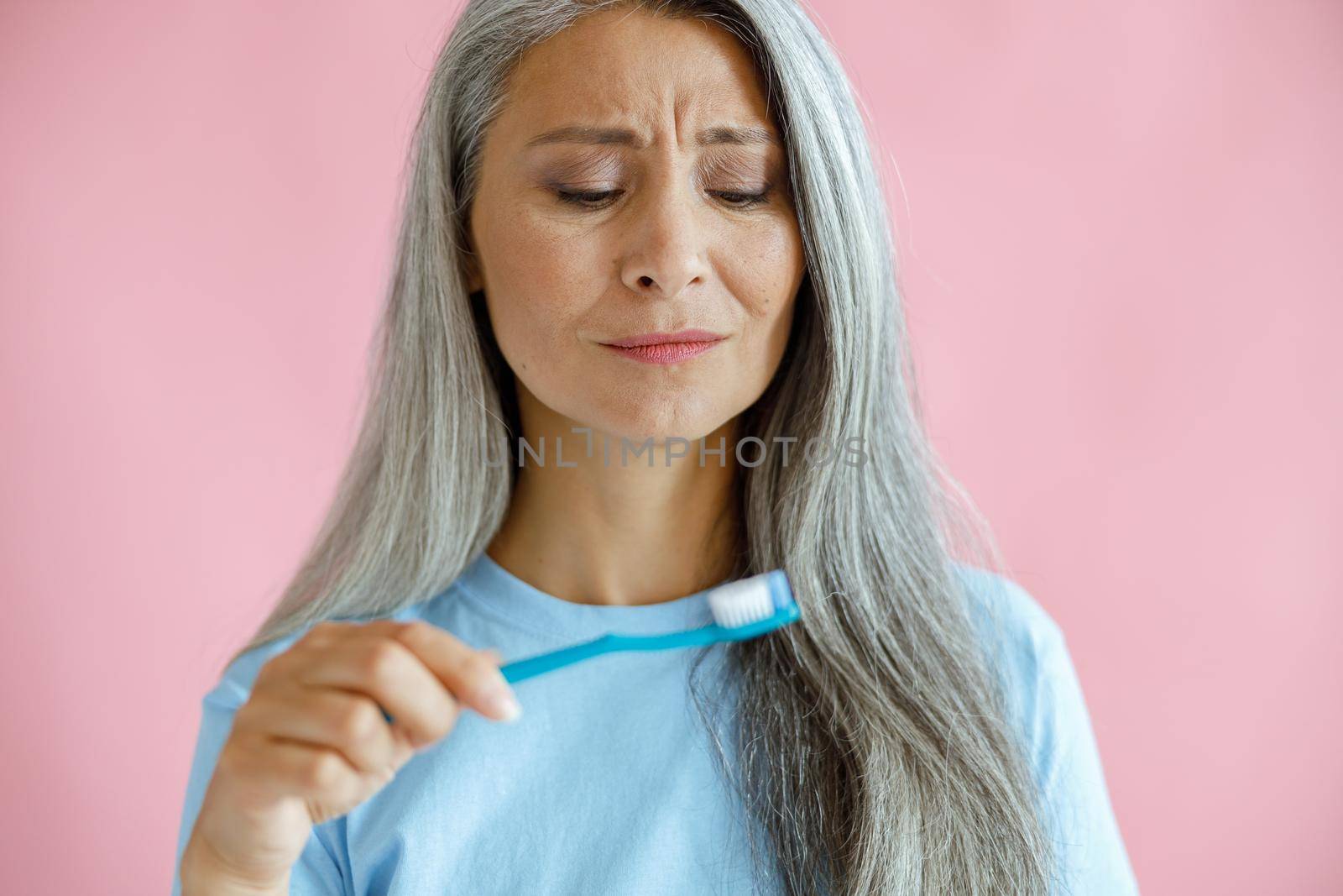 Upset mature Asian lady in blue t-shirt looks at toothbrush posing on pink background by Yaroslav_astakhov