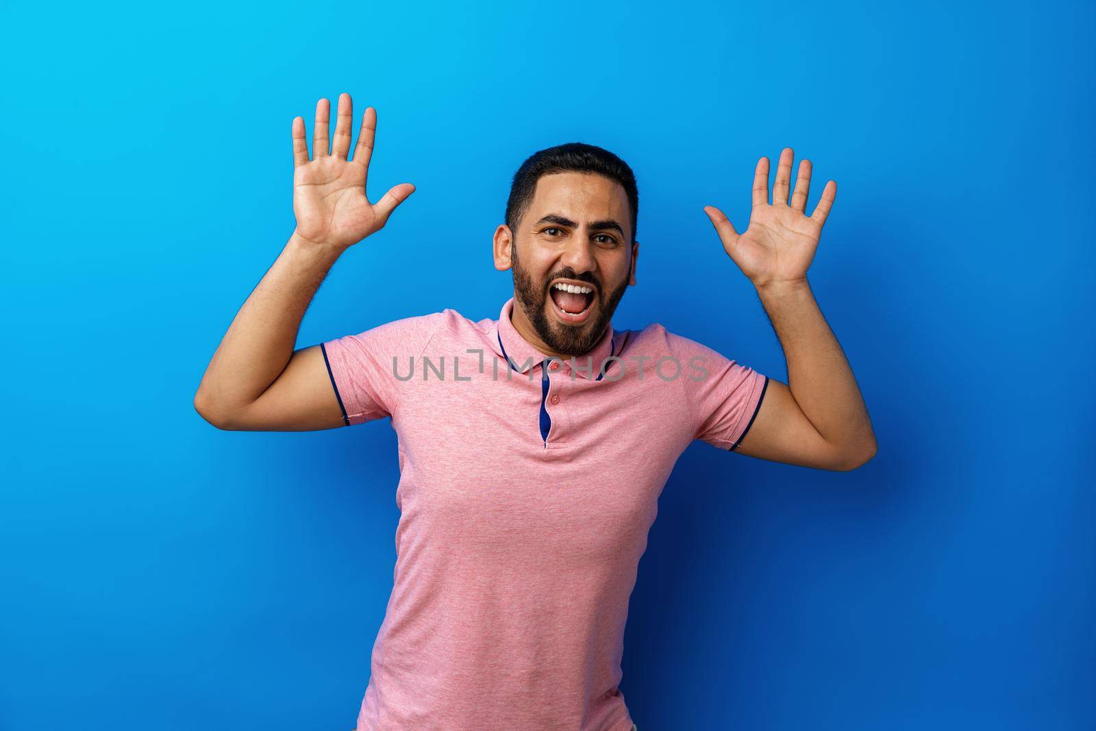 Portrait of amazed brunette man with beard in pink t-shirt against blue background, close up
