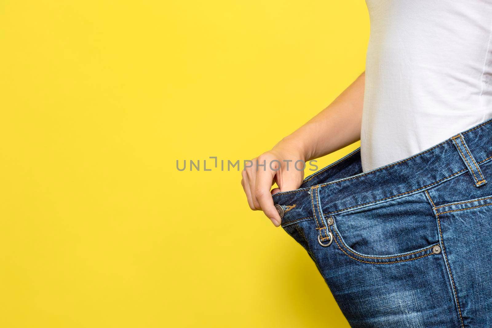 The concept of diet, proper nutrition, weight loss. Slim Woman Showing Loose Jeans and her Loss Weight. Woman white t-shirt and oversize jeans isolated on yellow background. Banner with copy space.