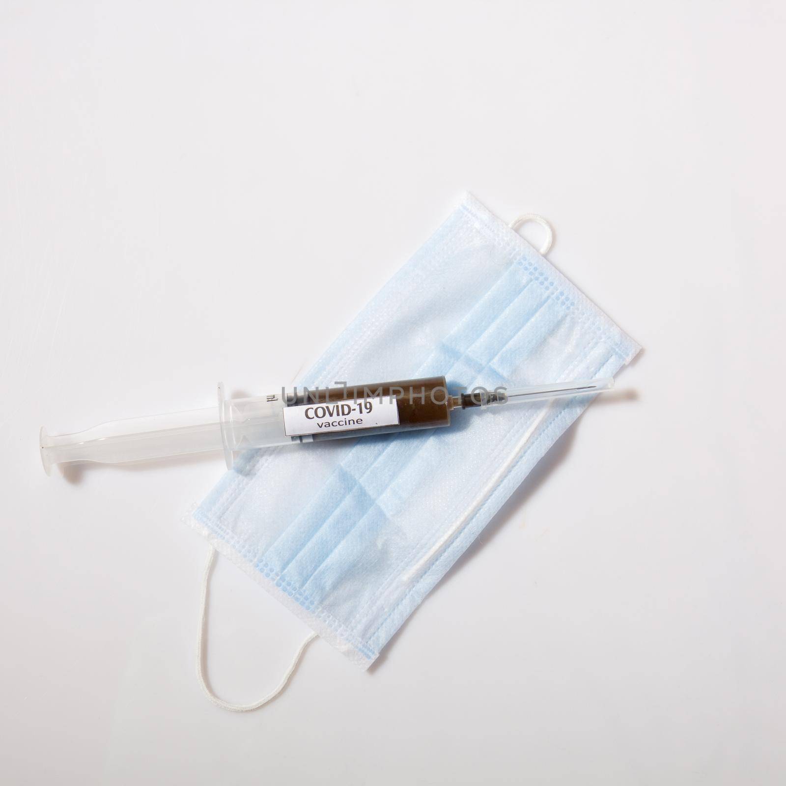Close up view of needle on a syringe filled with vaccine laying on face mask. on gray background