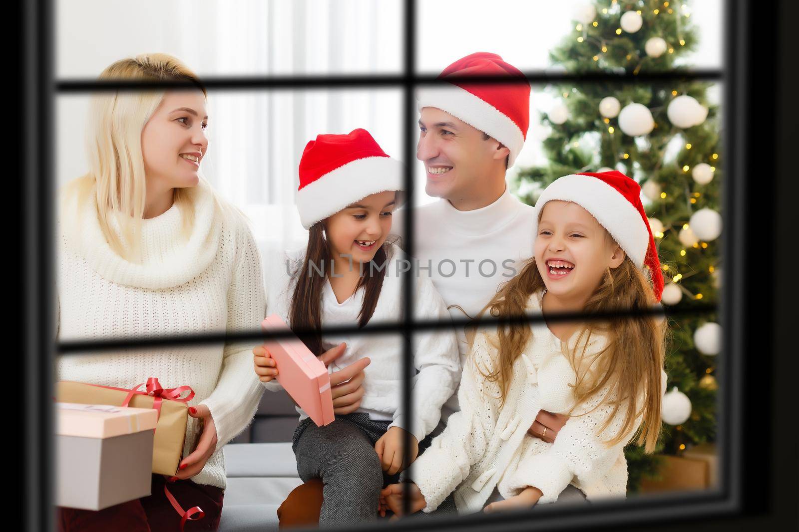 Young big family celebrating Christmas enjoying dinner, view from outside through a window into a decorated living room with tree and candle lights, happy parents eating with three kids